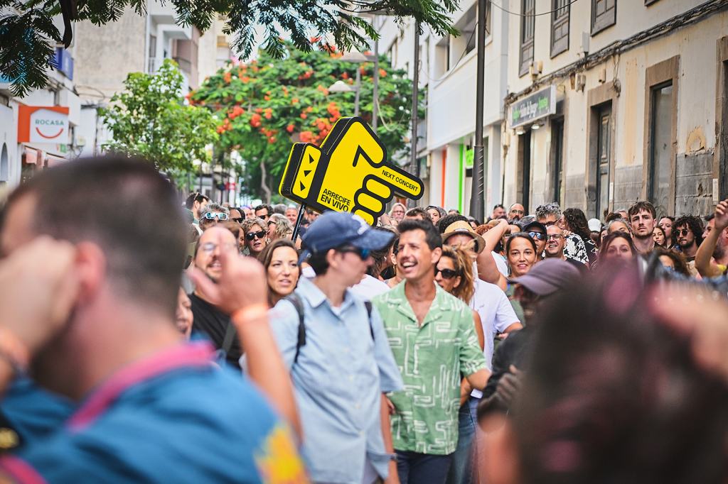 A lively crowd gathered in an urban street, with diverse attendees smiling and enjoying the atmosphere. A large yellow sign with a pointing hand promotes an upcoming concert.