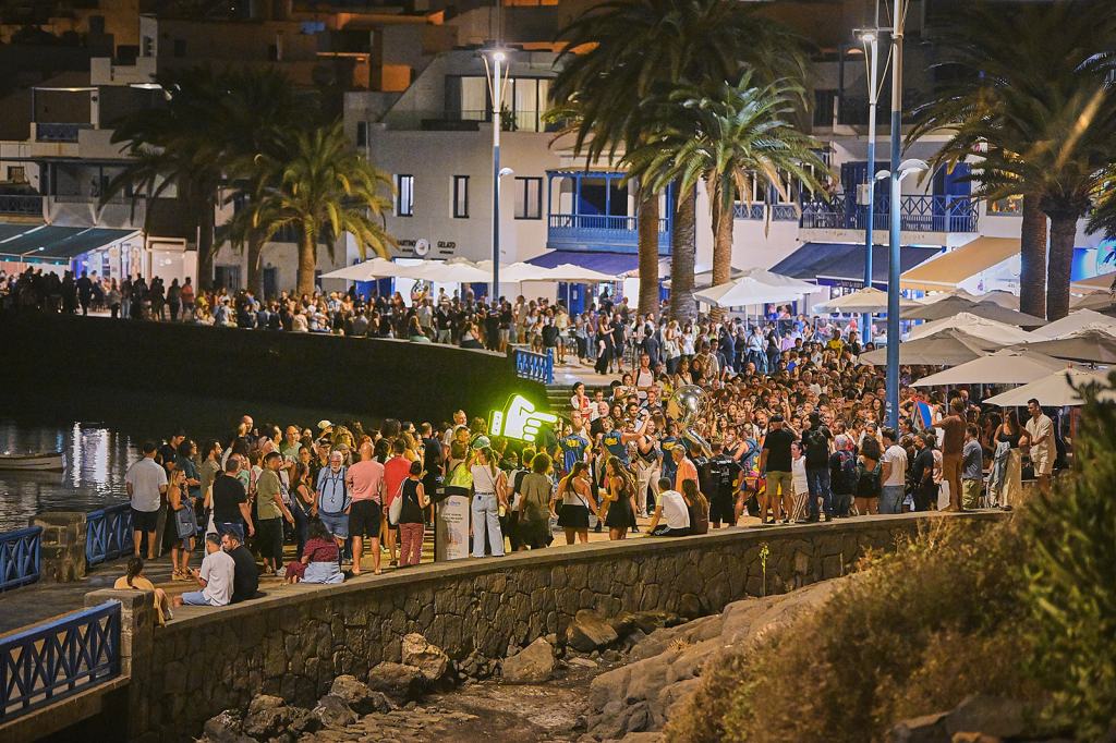 A large crowd gathered along a waterfront promenade in the evening, with palm trees and buildings in the background. People are socializing and enjoying the lively atmosphere.