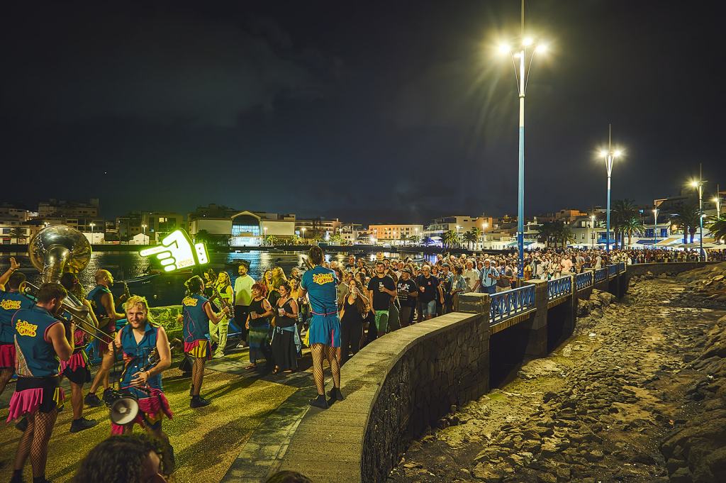 A nighttime scene depicting a vibrant crowd gathered along a waterfront promenade, with performers in colorful costumes playing instruments and a lit sign pointing towards the audience.