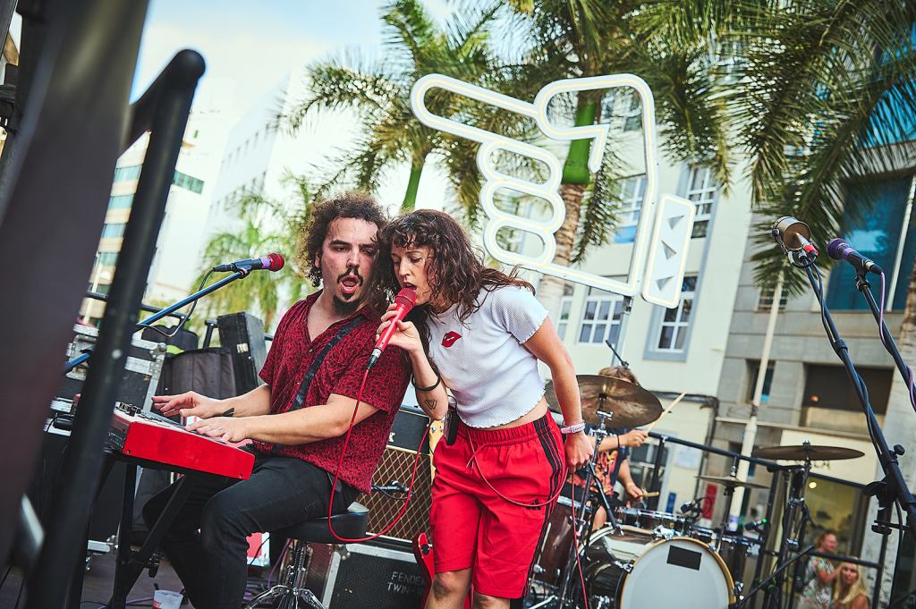A musician playing a red keyboard while another singer performs passionately on stage, surrounded by palm trees and a large thumbs-up sign in the background.