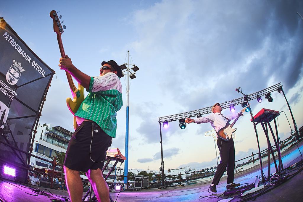 Two musicians performing on stage at an outdoor concert, one playing a green bass guitar and the other a guitar, with colorful stage lights and a cloudy sky in the background.