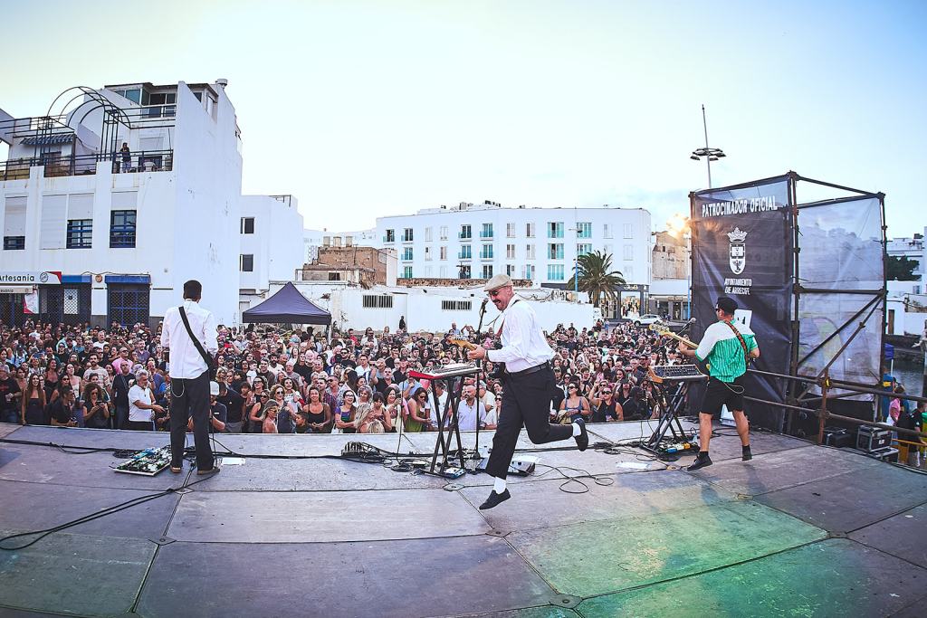 A lively music performance on an outdoor stage with a large crowd in attendance, featuring three musicians playing instruments and engaging with the audience against a backdrop of white buildings and palm trees.