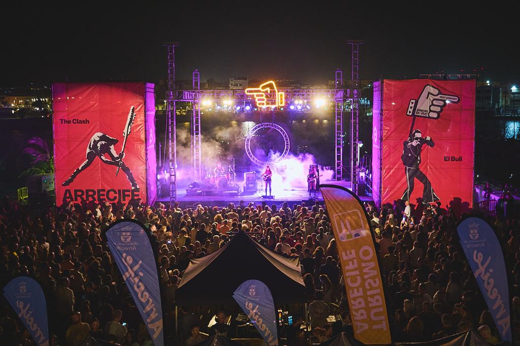 A lively outdoor concert scene at night with a large crowd of audience members in front of a stage. The stage is lit with colorful lights, surrounded by large banners featuring artistic designs. Two prominent banners display the names "The Clash" and "El Bulli." Smoke effects add to the dynamic atmosphere.