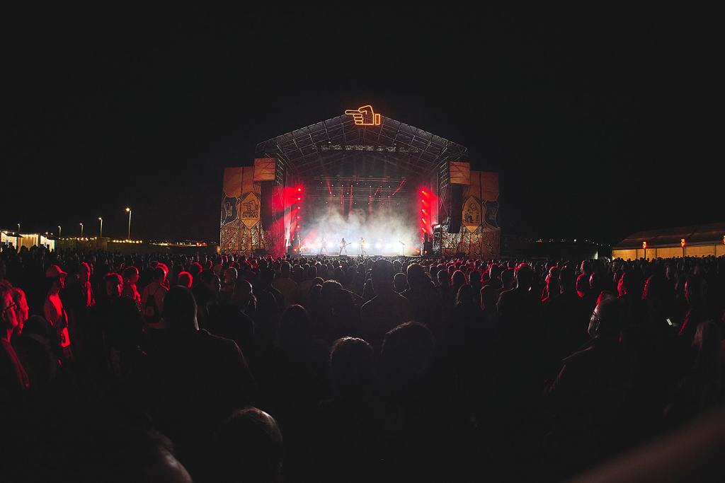 A large crowd at a nighttime outdoor music festival, illuminated by red lights from the stage where performers are visible.