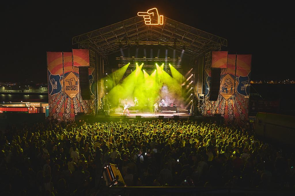 A large crowd at a nighttime concert with a brightly lit stage, featuring musicians performing under colorful lights and banners.