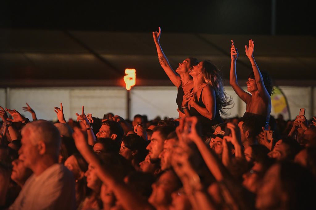 A lively crowd at a concert, with people cheering and raising their hands in excitement. Some individuals are capturing the moment on their phones, while a few are close together, enjoying the performance.